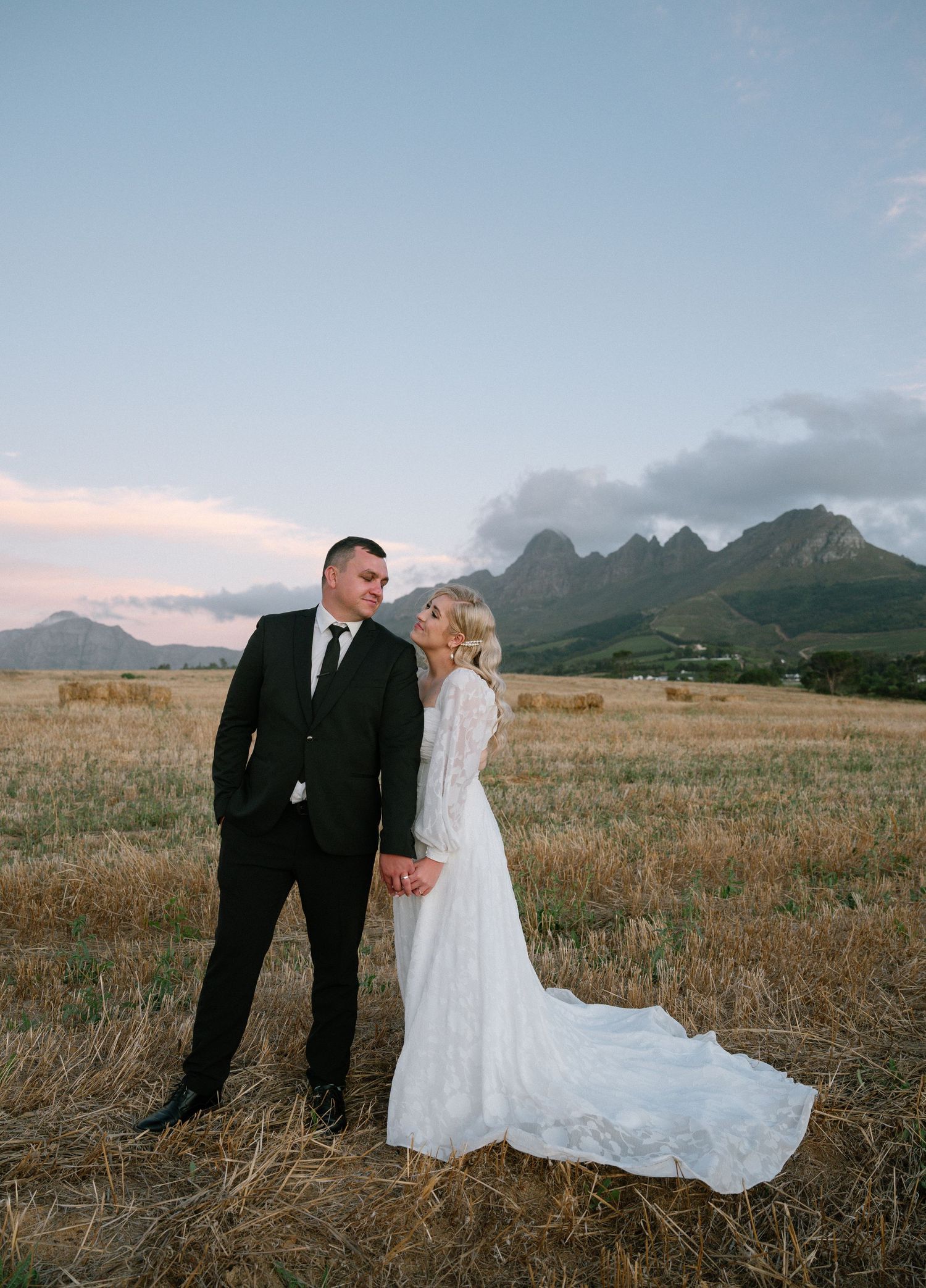 A wedding couple poses in a field with dramatic mountain peaks in the background.