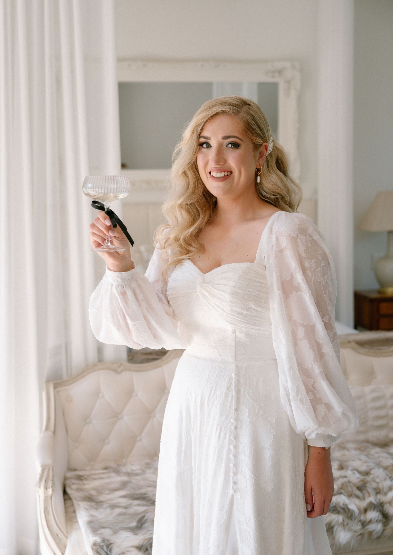 A person wearing a romantic white wedding dress with puff sleeves stands holding a champagne glass in an elegant room.