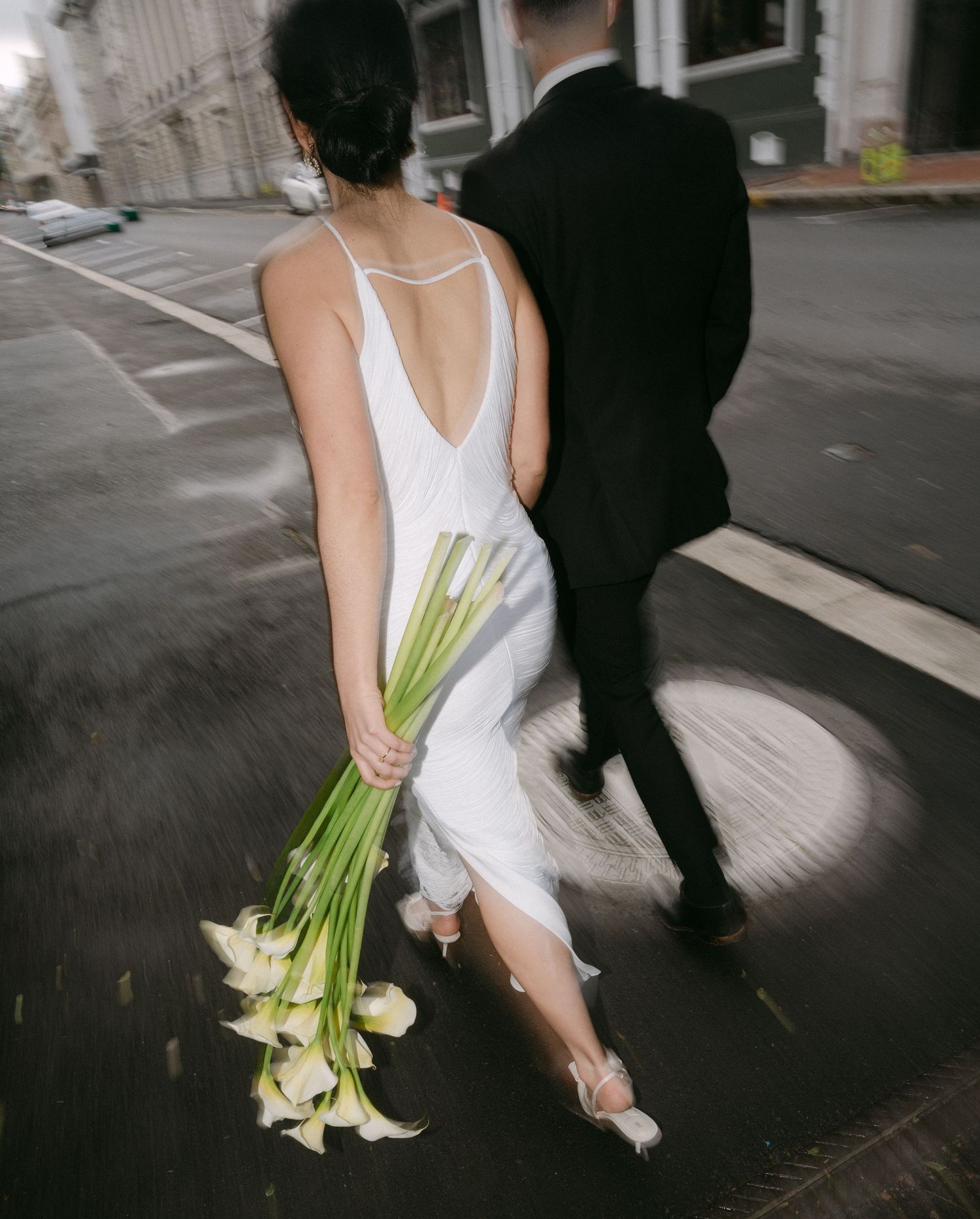 Cropped wedding photo shows bride in white dress and groom in black suit walking with calla lilies.