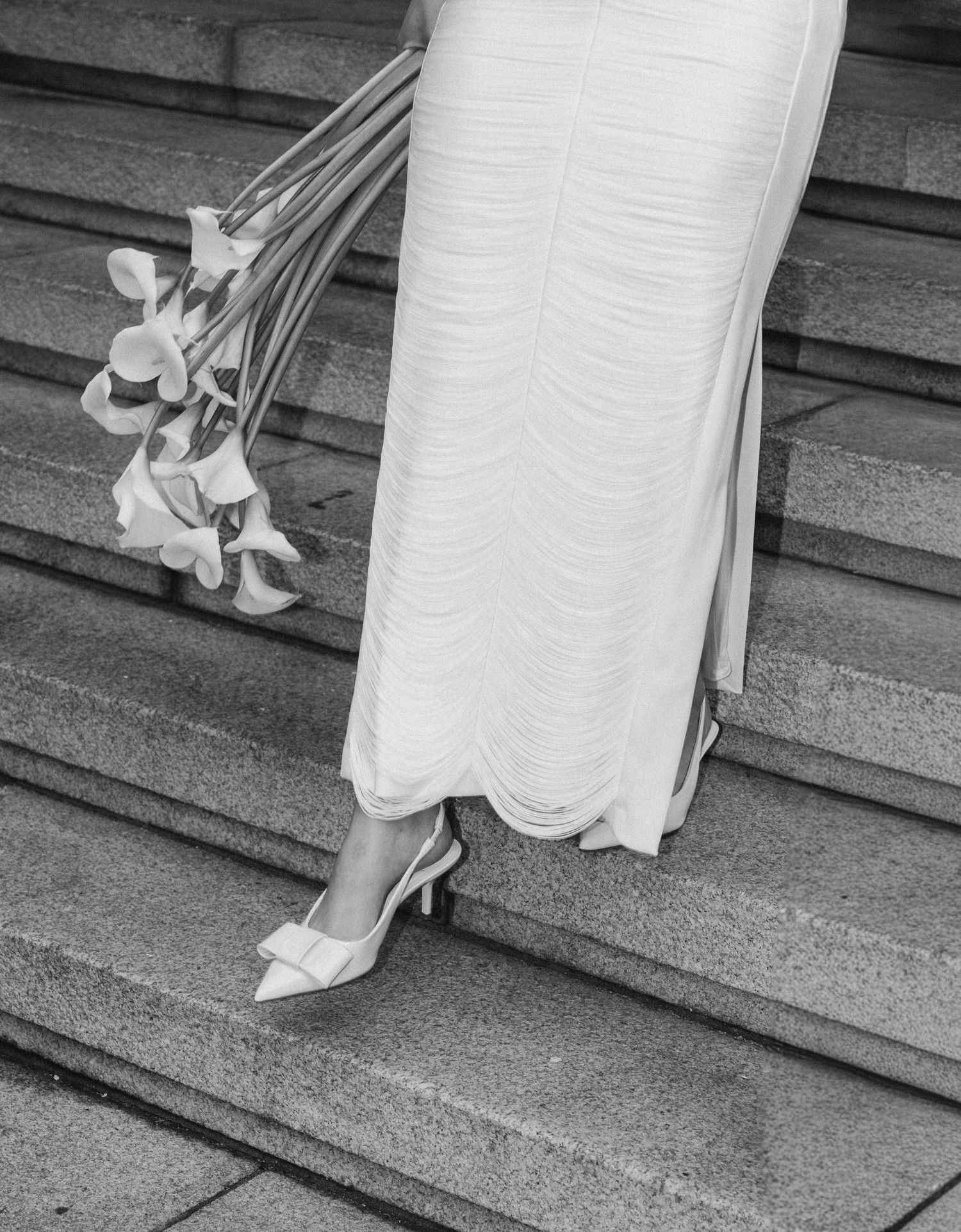 Black and white image showing a person in a white dress ascending stone stairs while holding onto a railing.