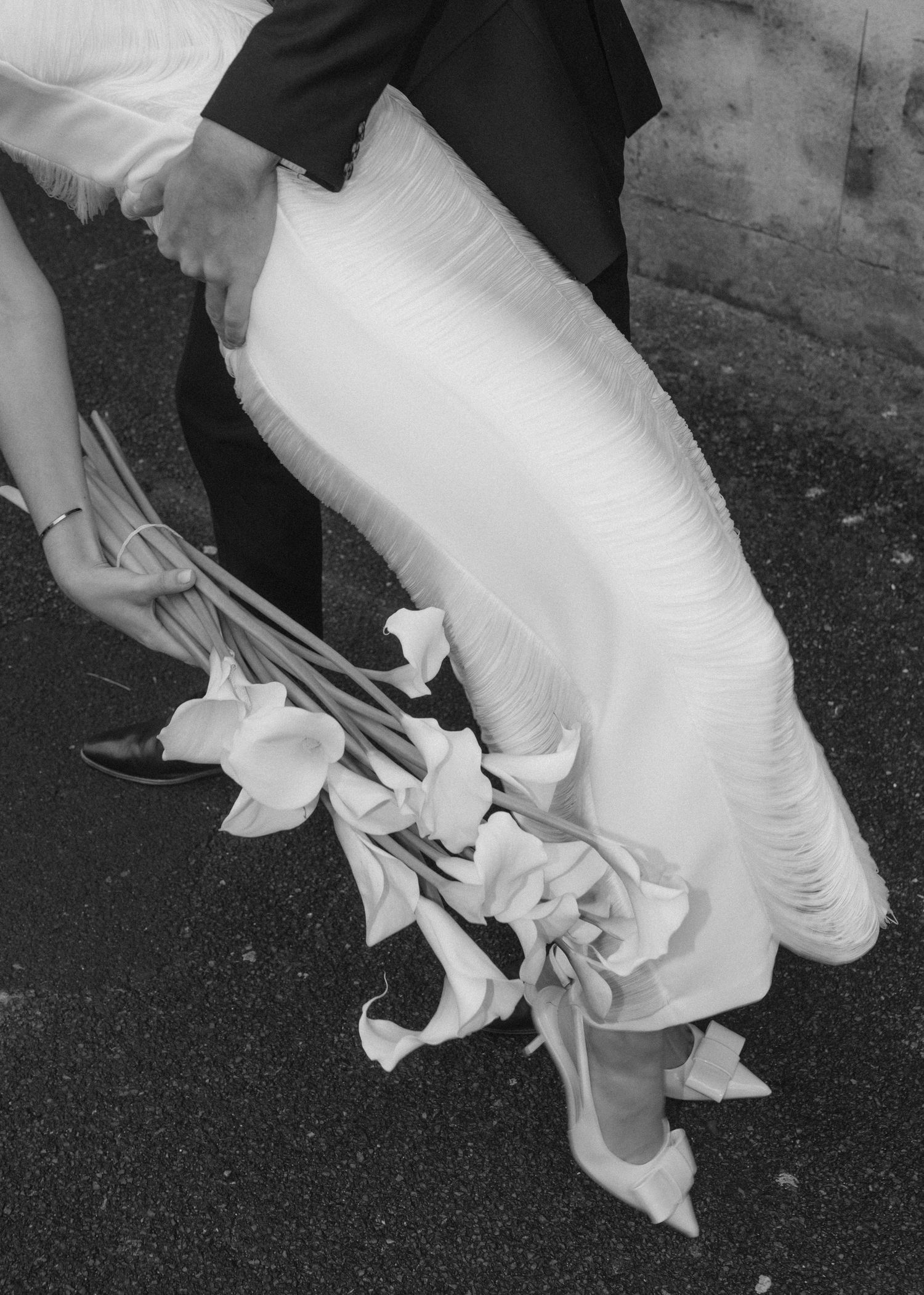 Black and white wedding photo showing elegant white dress and bouquet of calla lilies against dark ground.
