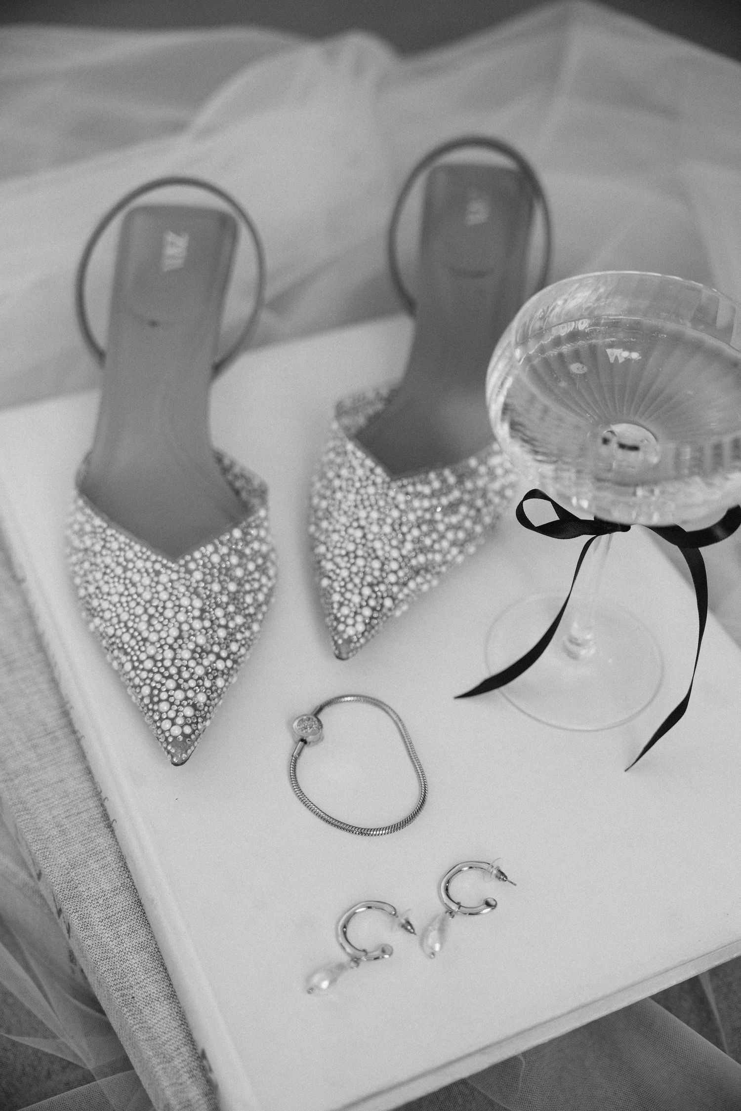 Sparkly bridal shoes, jewelry and a champagne glass arranged on white tulle fabric.