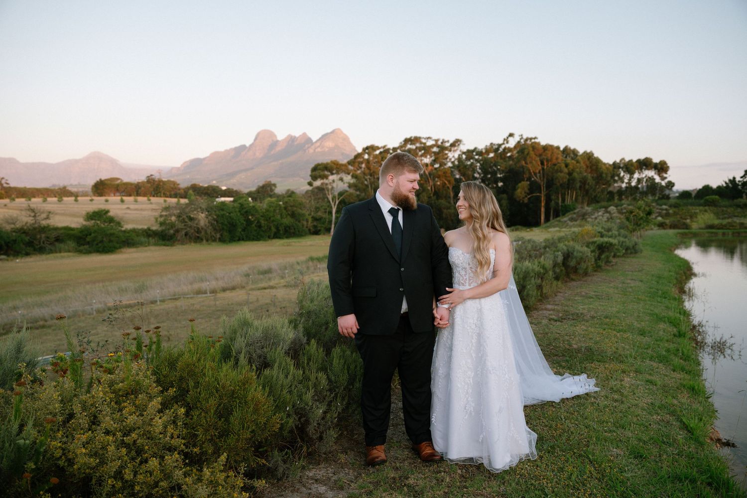 Wedding couple stands by serene lake with mountain backdrop at sunset.