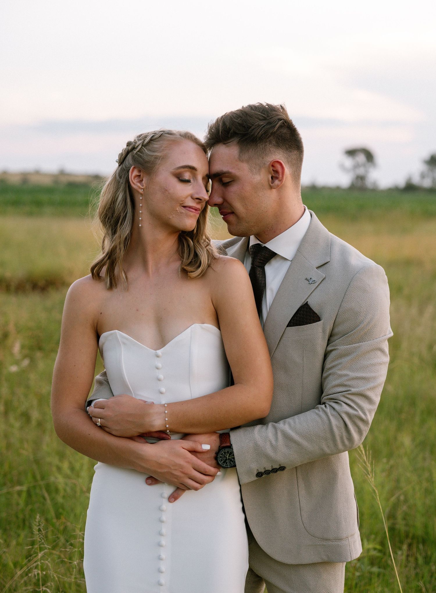 A romantic wedding couple embraces in a scenic field during golden hour.