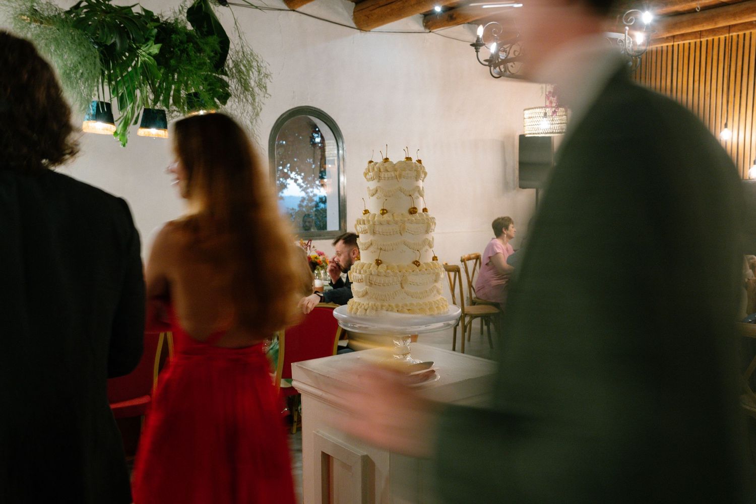 A white wedding cake displayed at a reception venue with guests in formal attire in the background.