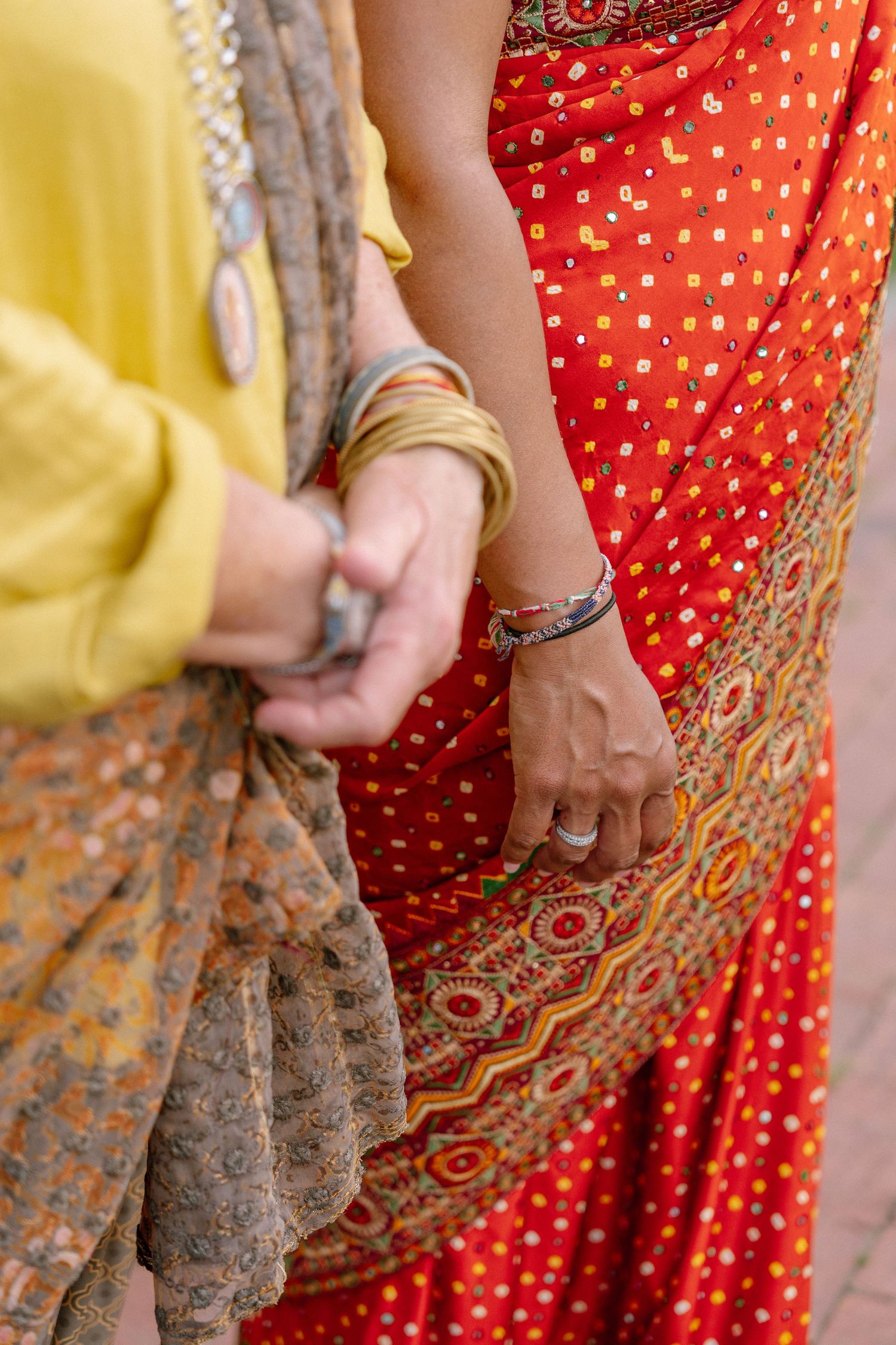 Close-up view of hands wearing bangles against traditional Indian saris in red and brown with gold embellishments.