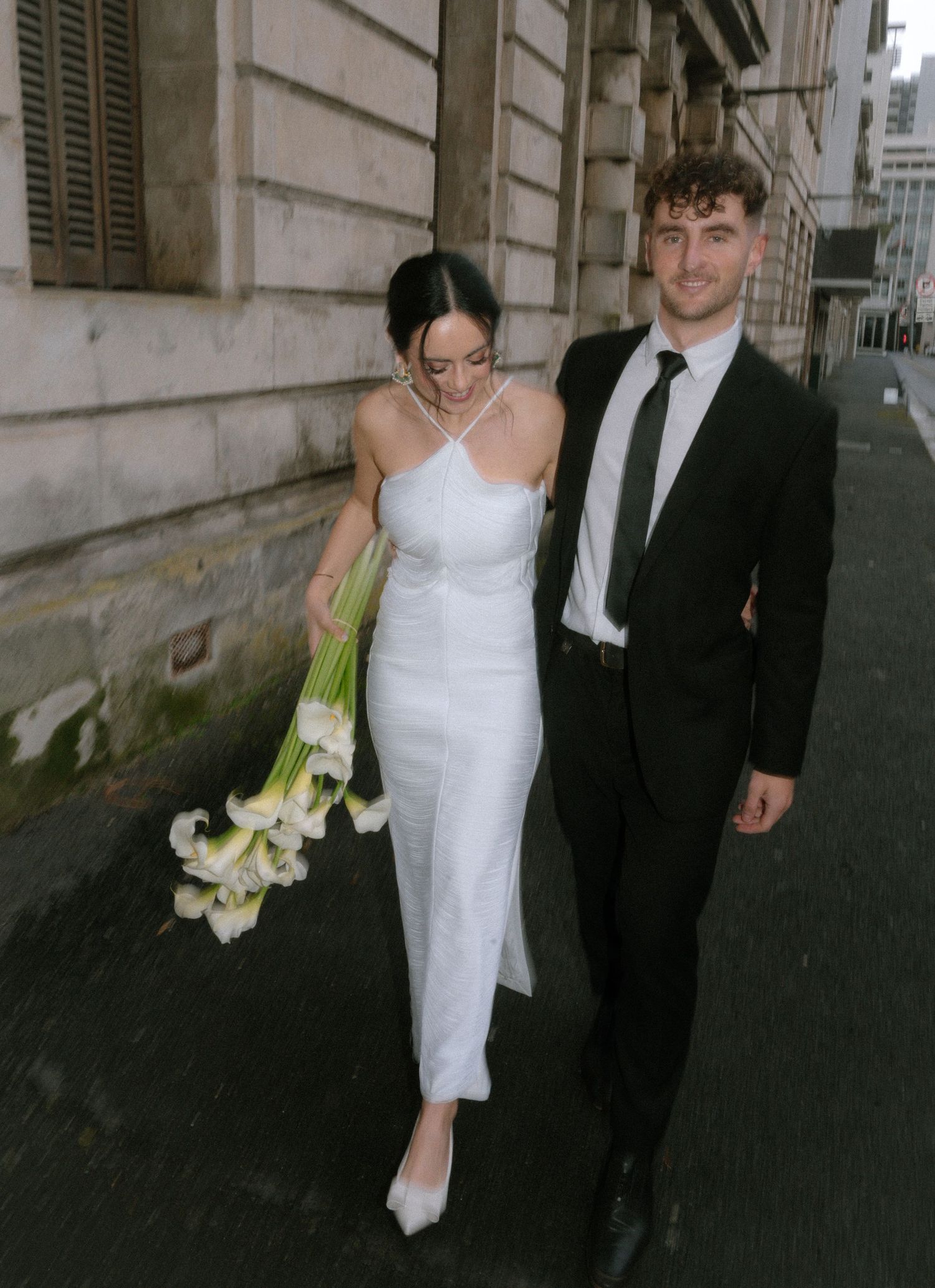 A couple walks down a city street, the person in white carrying a bouquet of white calla lilies.