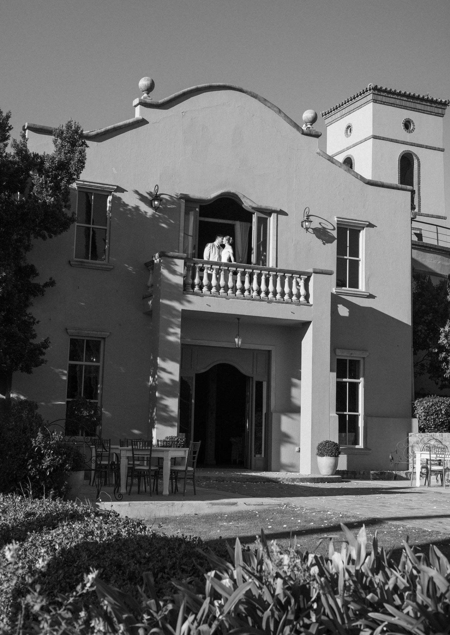 Black and white exterior view of a Mediterranean-style villa with white walls and arched doorways surrounded by landscaping.