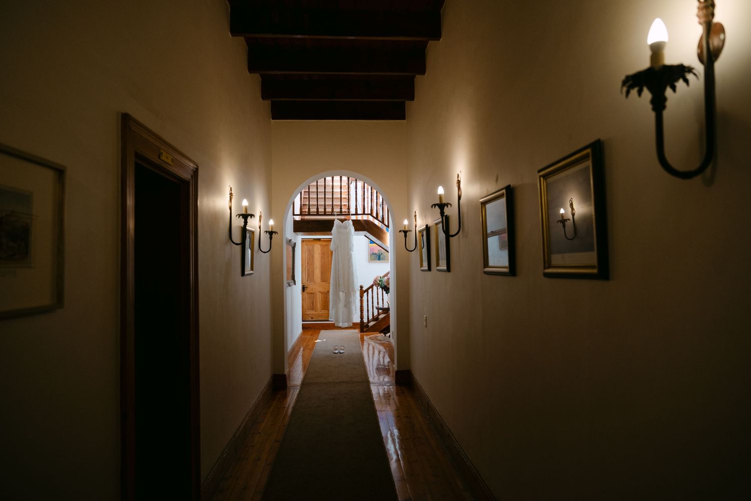 A dimly lit hallway with arched doorway and wall sconces casting warm light on beige walls and hardwood floors.