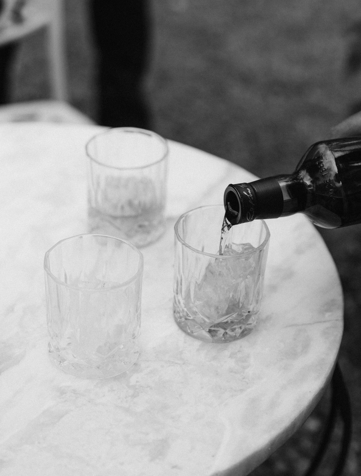 Black and white photo of wine being poured into crystal glasses on a white marble table.