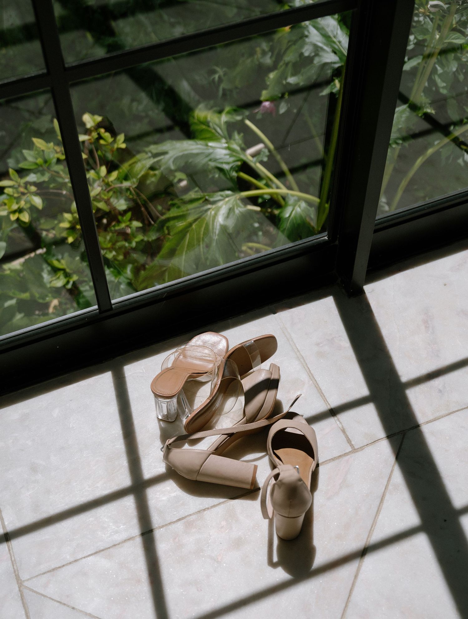 A pair of elegant beige high-heeled sandals sits on a sunlit floor by a window, casting dramatic shadows through the panes.