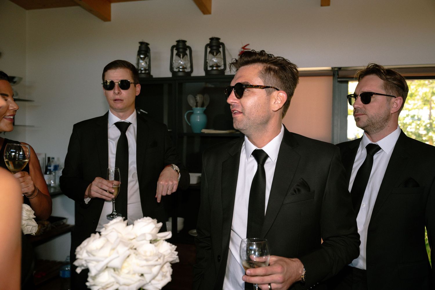 Three people in matching black suits and sunglasses holding drinks at a formal event.
