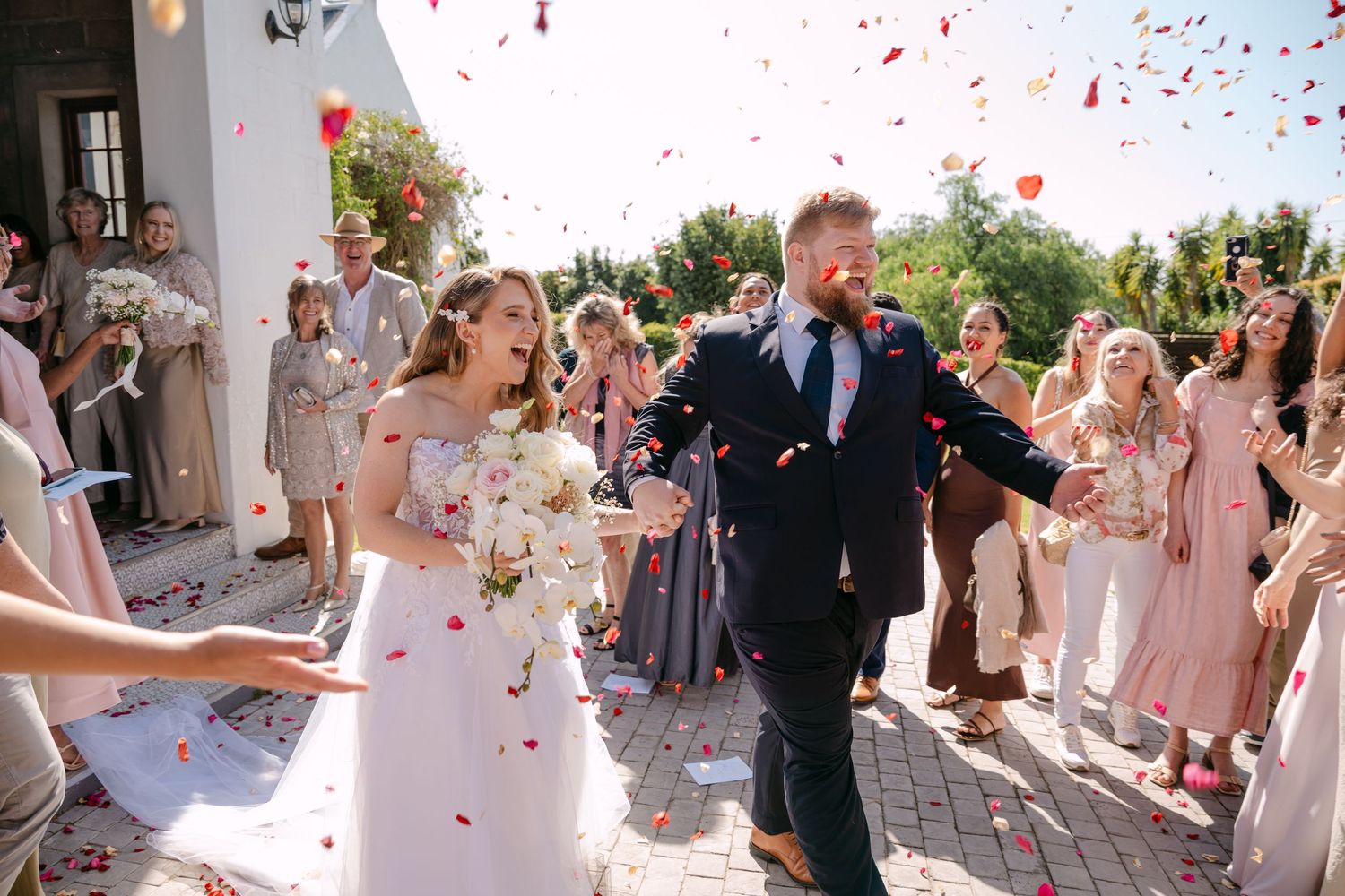 Newlyweds exit ceremony while guests shower them with rose petals on sunny day.