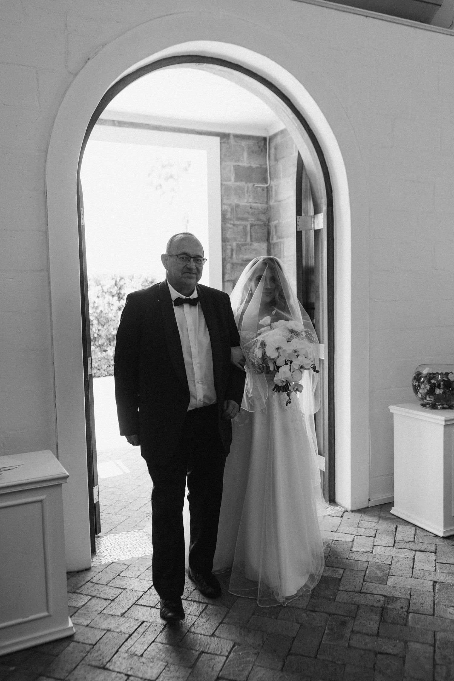 Black and white photo of bride and escort walking through arched doorway.
