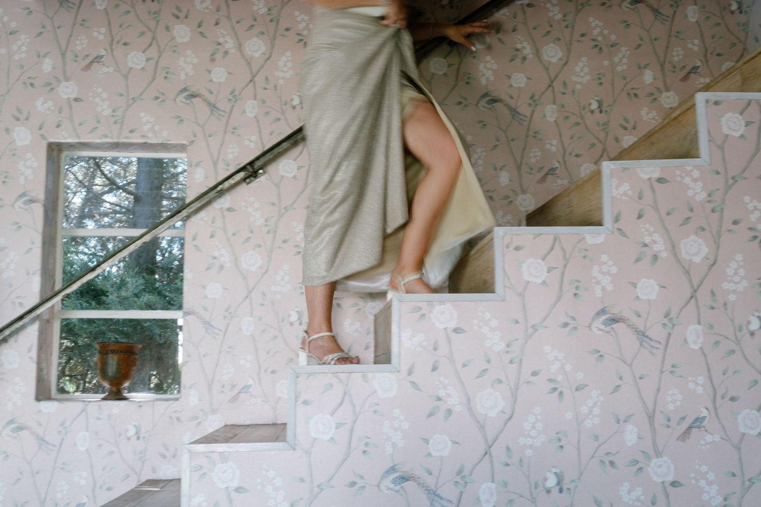 Person ascending white stairs against floral wallpaper with a window visible in the corner.