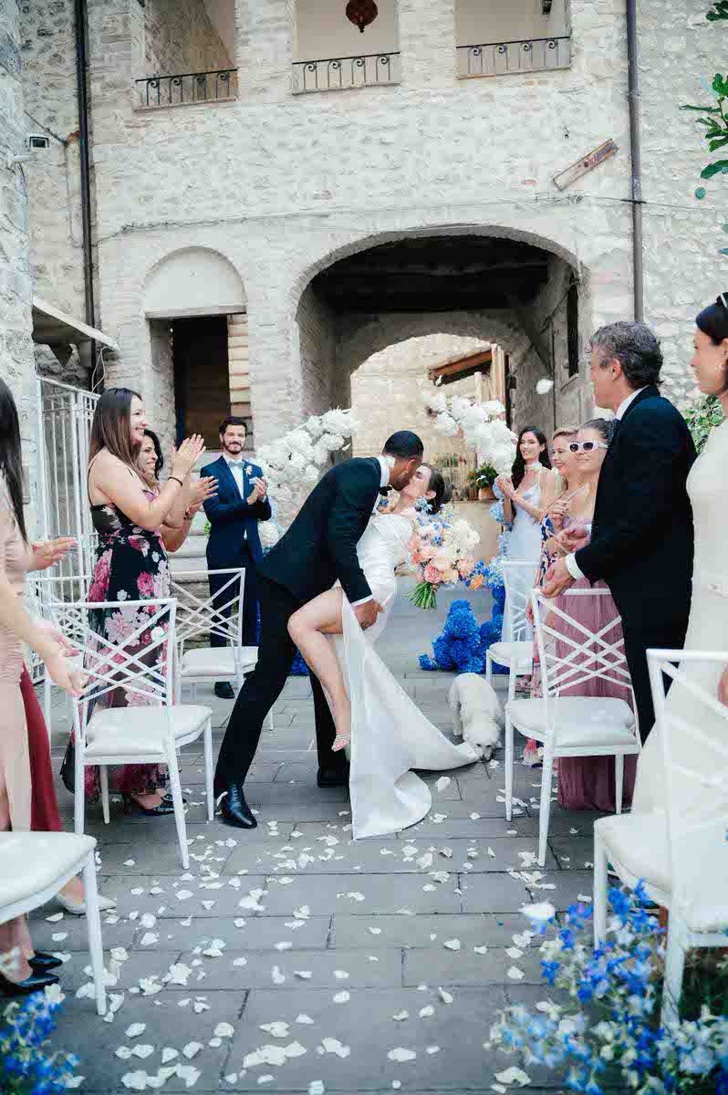 Wedding ceremony moment in stone courtyard decorated with blue flowers and white chairs.