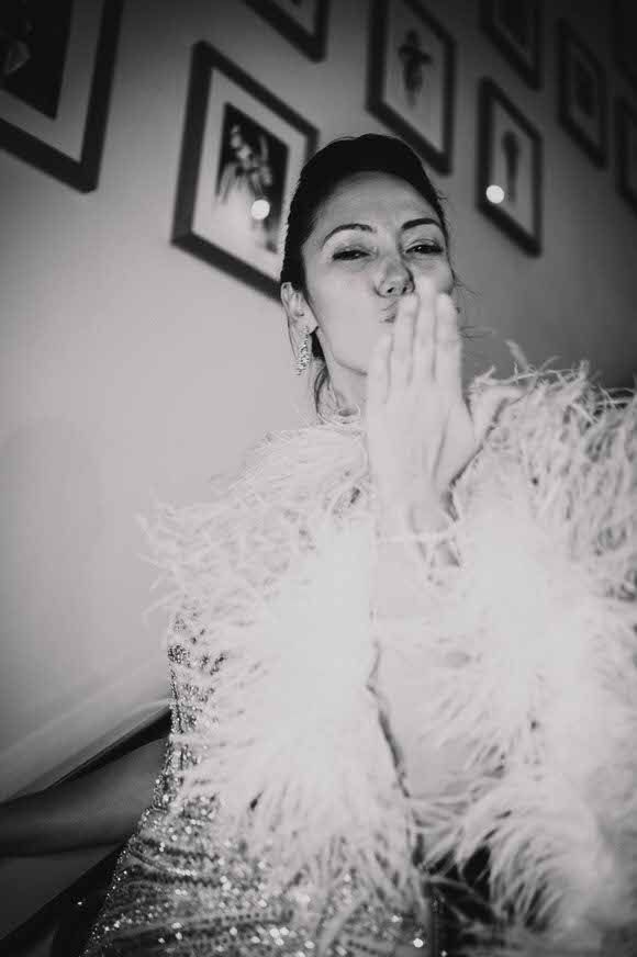 Black and white photo of a glamorous woman in feathered outfit blowing a kiss, framed pictures on wall behind her.