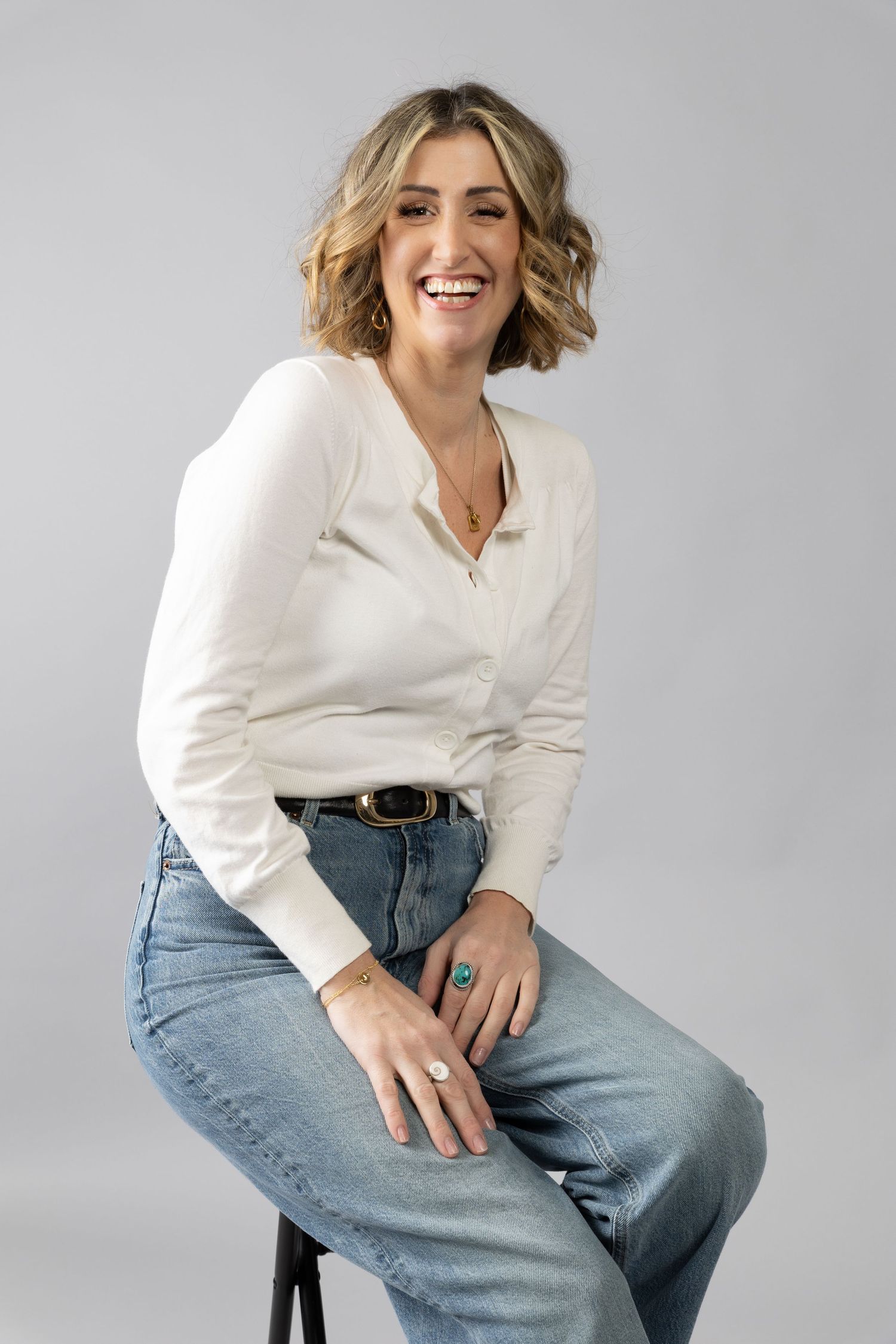 Casual studio portrait featuring a white blouse, jeans and wavy blonde hair against a grey background.