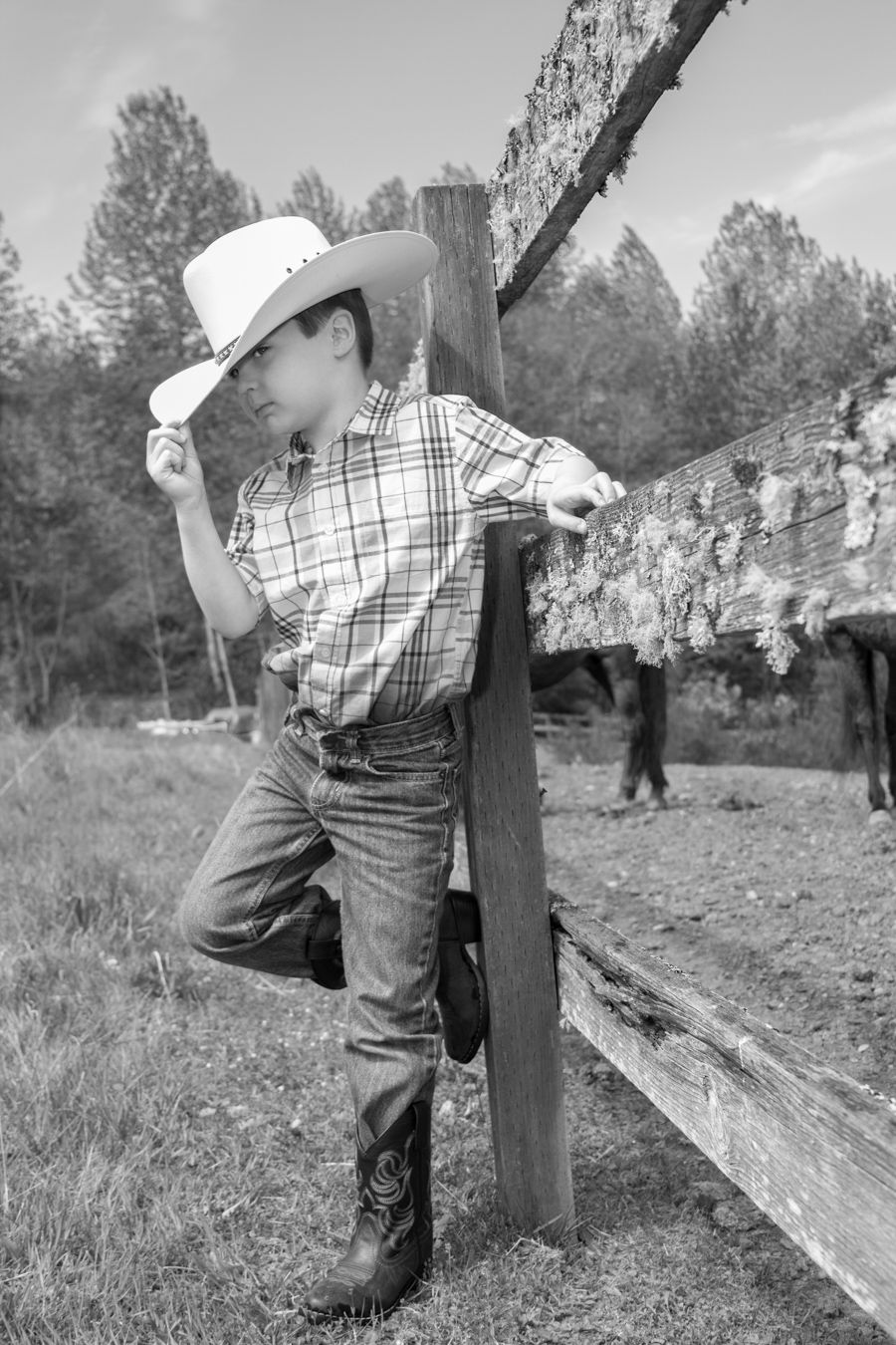 Black and white photo of young cowboy leaning on wooden fence post in rustic setting.