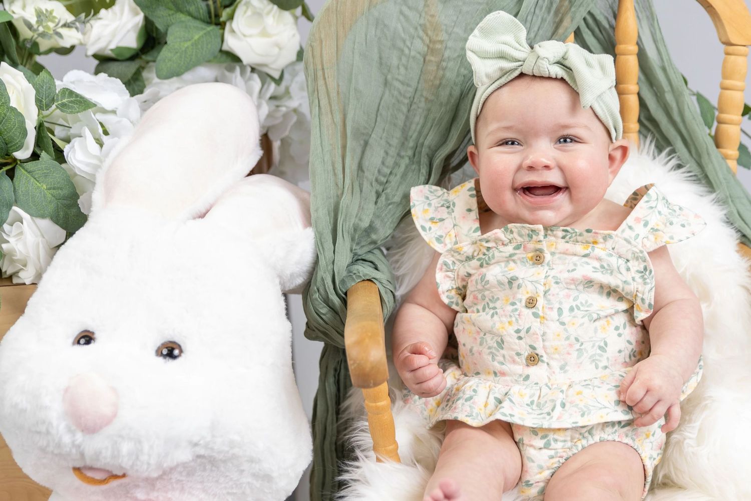 Baby in floral outfit lying on white bedding with green foliage accents.