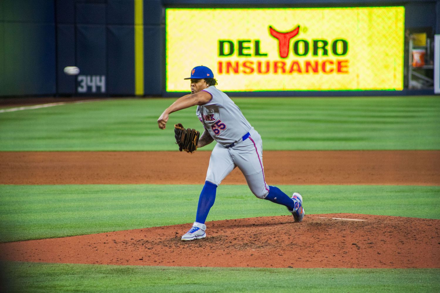 Baseball pitcher in blue and white uniform throws pitch at night game with Del Toro Insurance sign visible.