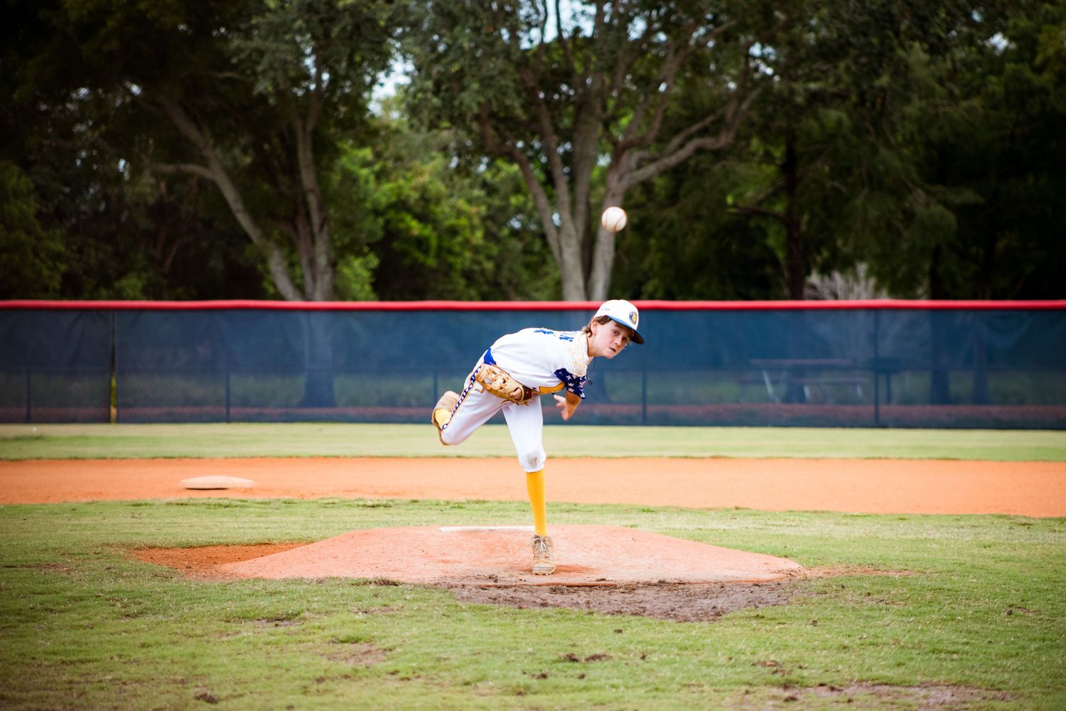 Baseball pitcher in white jersey and yellow socks follows through on delivery from pitching mound during game.
