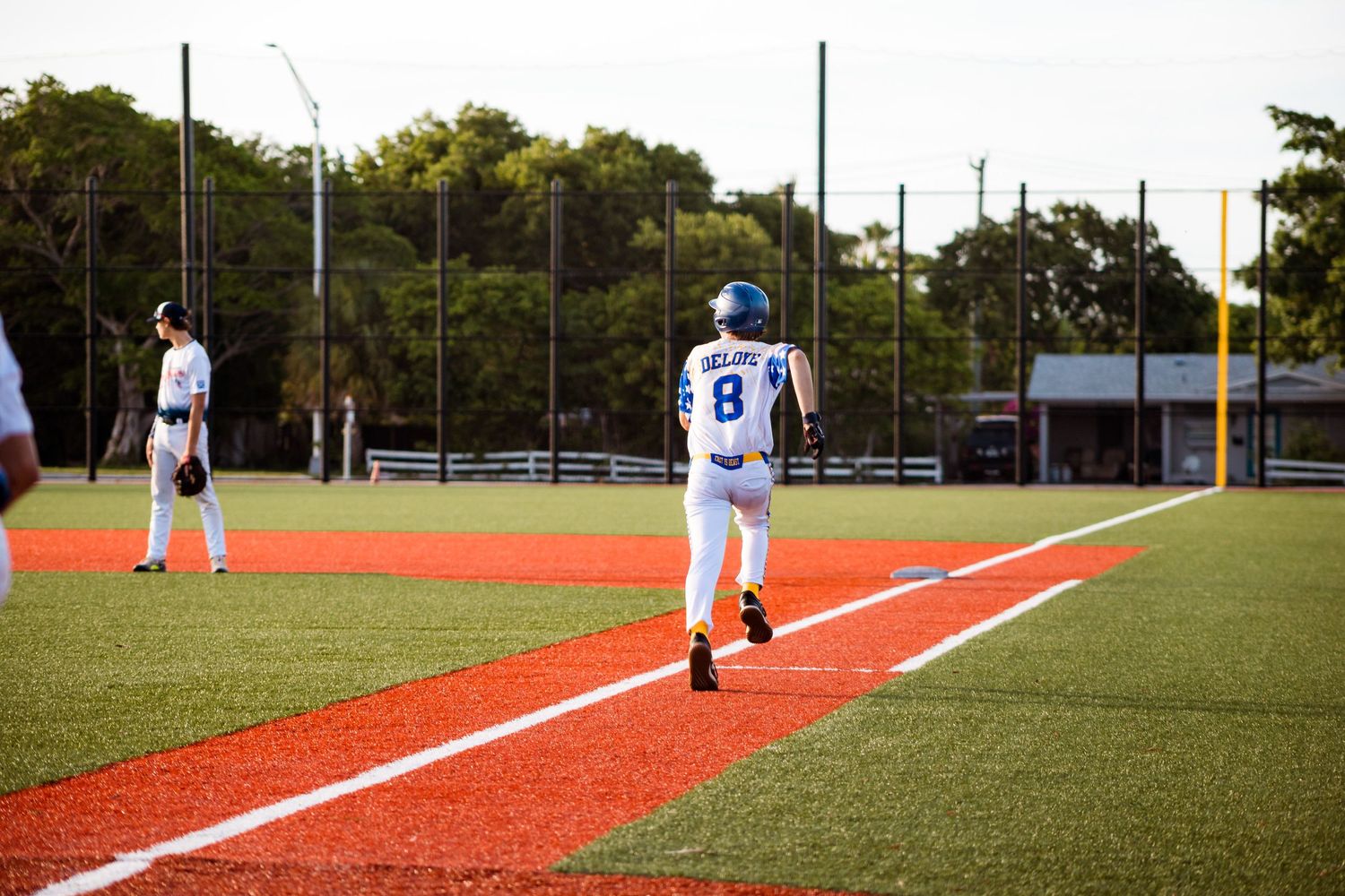 Baseball players in white uniforms stand on a bright red baseline of a baseball field during a sunny day.