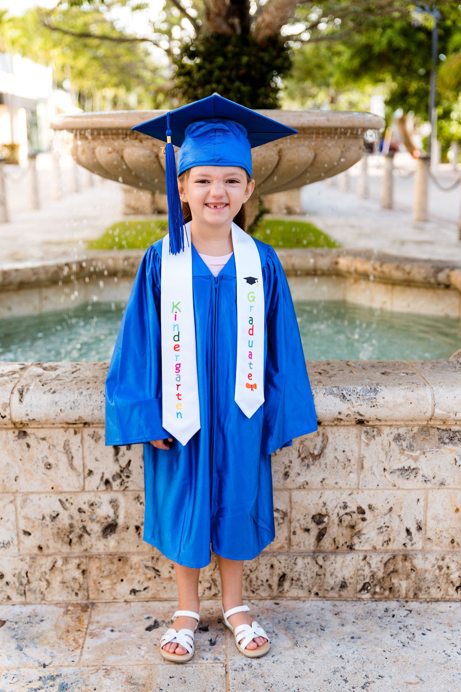 Young graduate wearing bright blue cap and gown stands smiling by fountain during kindergarten graduation ceremony.