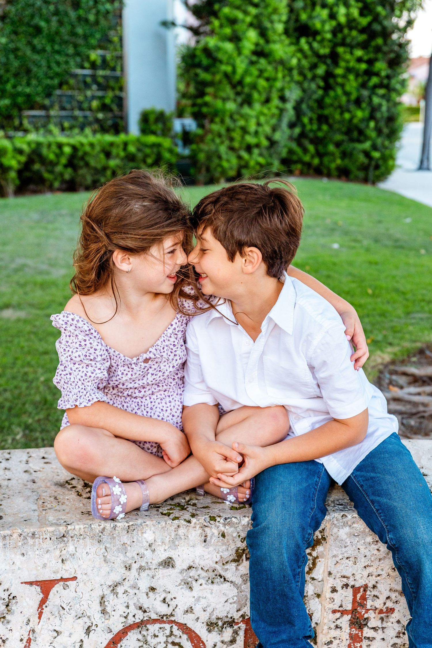 Two children sitting on a stone ledge touching foreheads and smiling, surrounded by lush green garden.