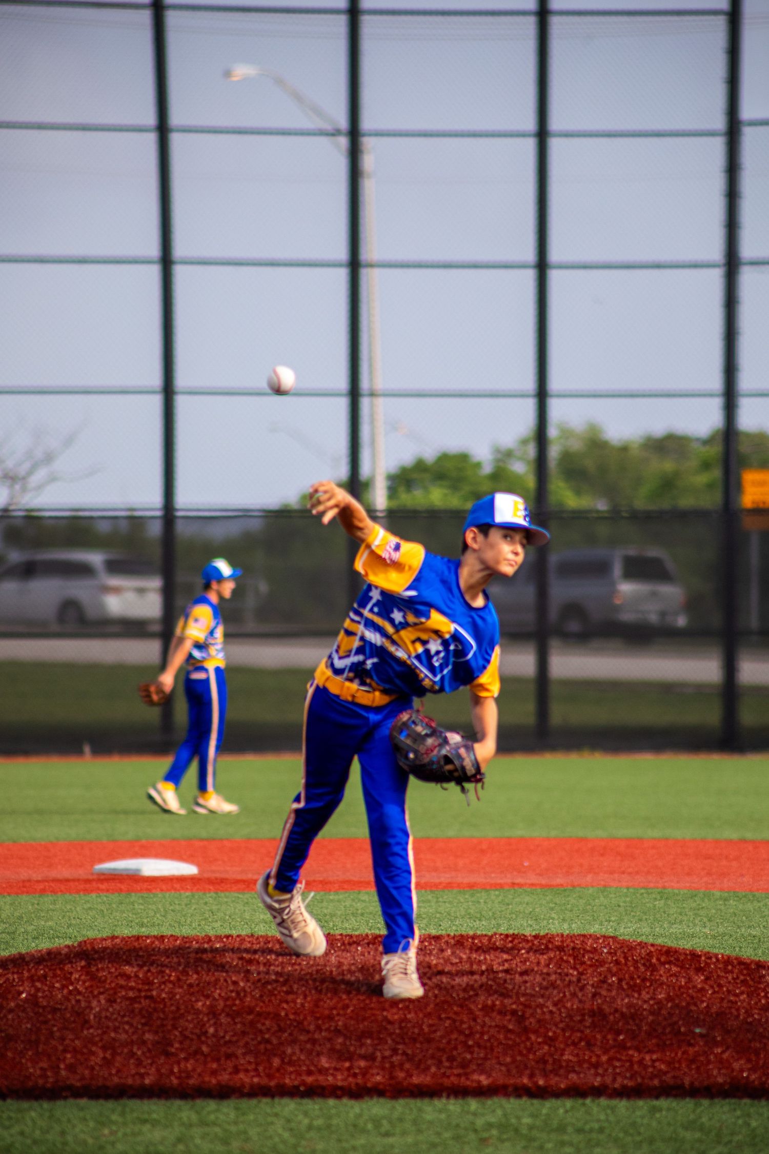 Young baseball pitcher in blue and gold uniform throwing a pitch from the mound during a youth game.