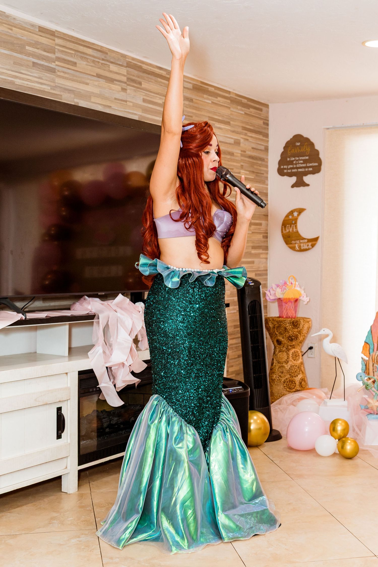 Performer in mermaid costume with sparkly green tail singing and raising arm during indoor party entertainment.