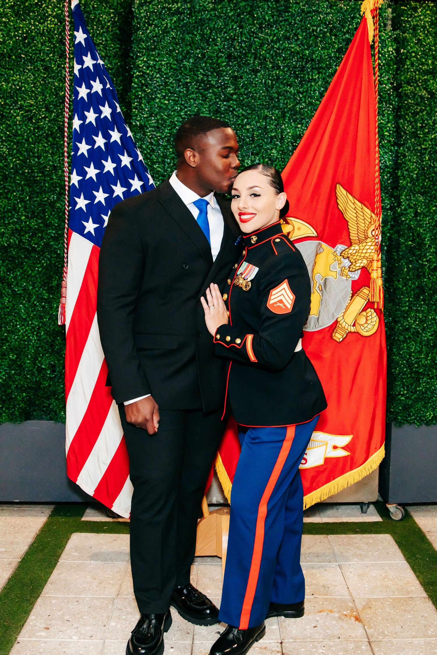 A couple in formal military attire poses together against a green hedge backdrop with American and Marine Corps flags.
