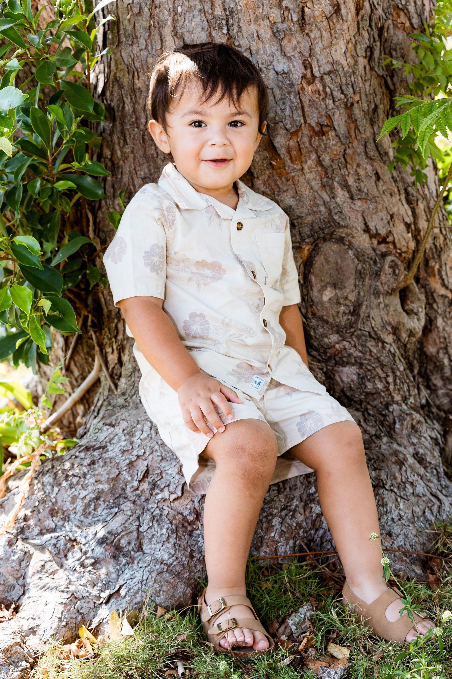 Child in white outfit sitting on large tree trunk in sunny garden setting.
