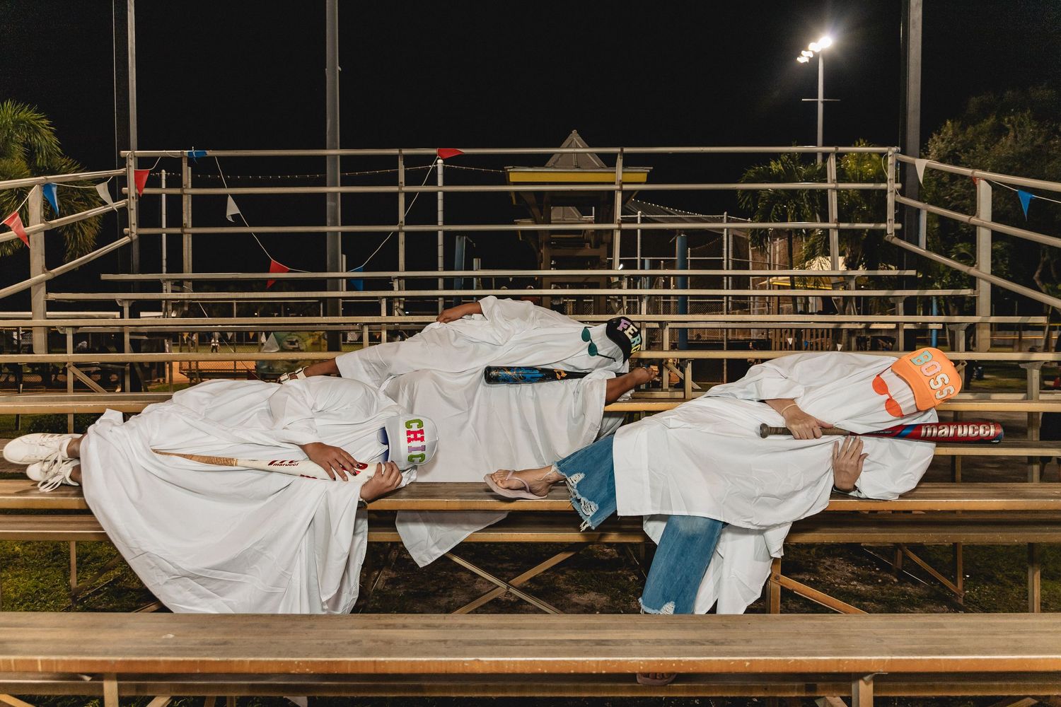 Row of people in white sheets doing synchronized movements on stadium bleachers at night.