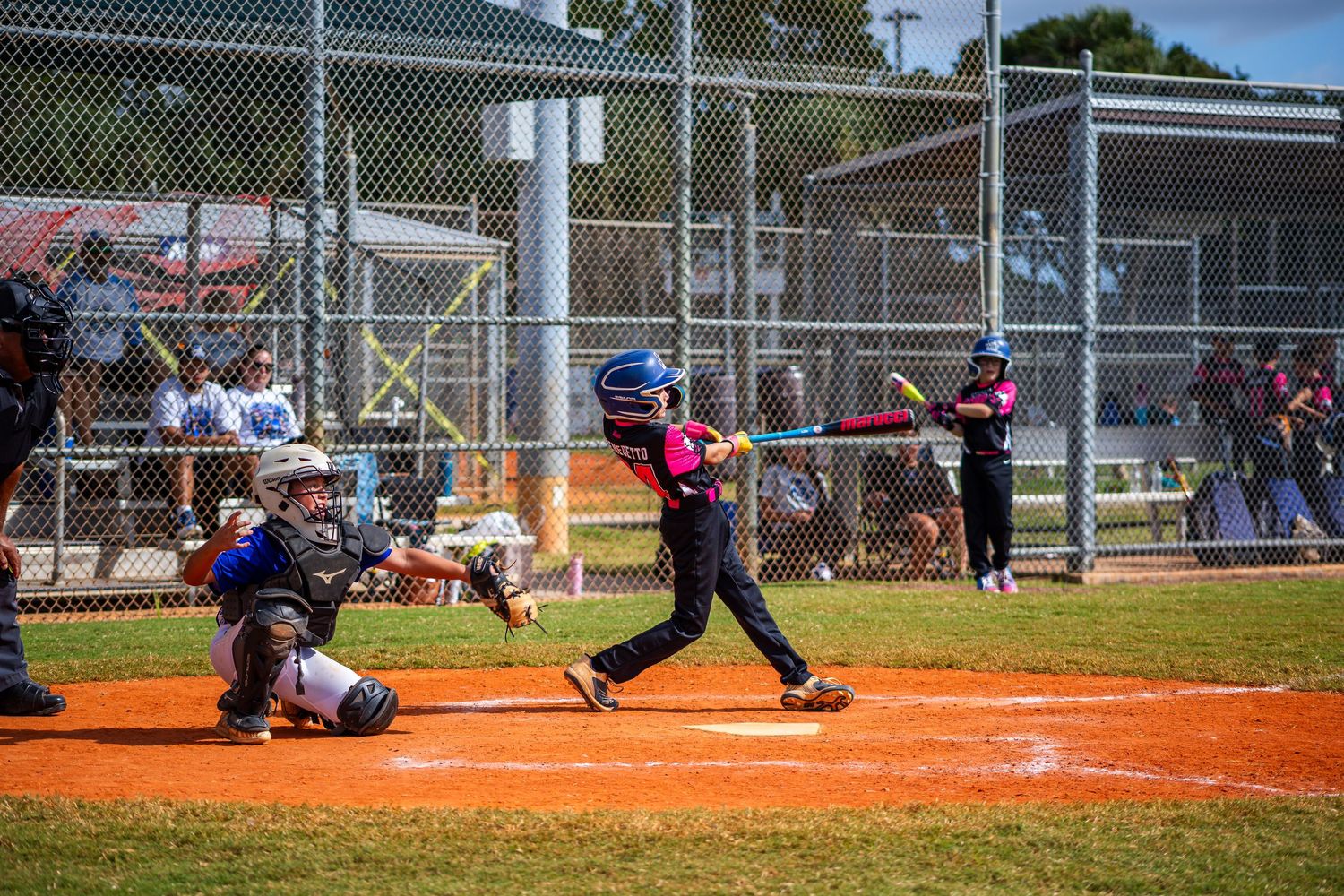 Youth baseball batter in purple uniform swings at pitch while catcher crouches behind home plate on dirt diamond.