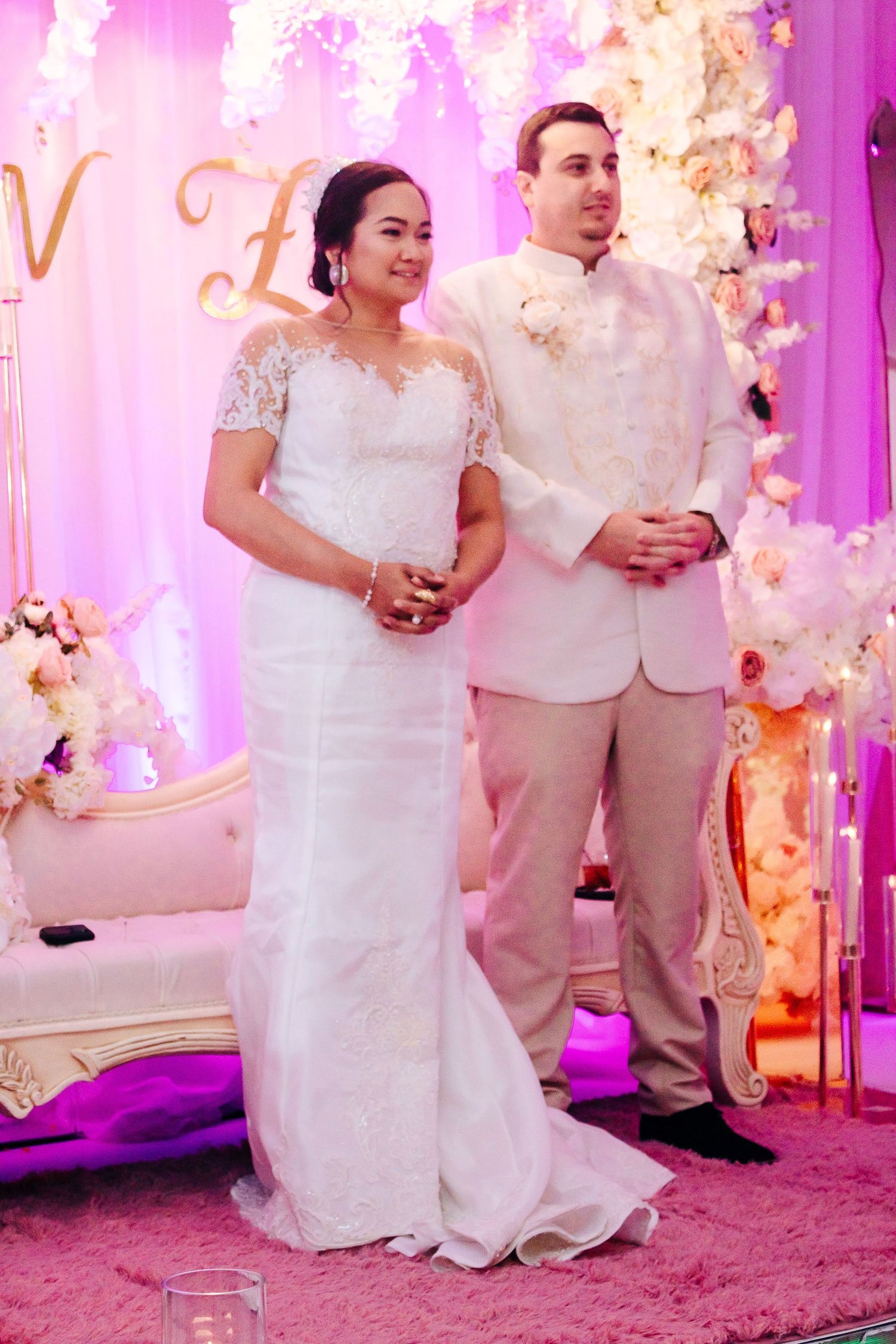Couple in white attire posing in front of pink floral wedding backdrop with decorative lighting.