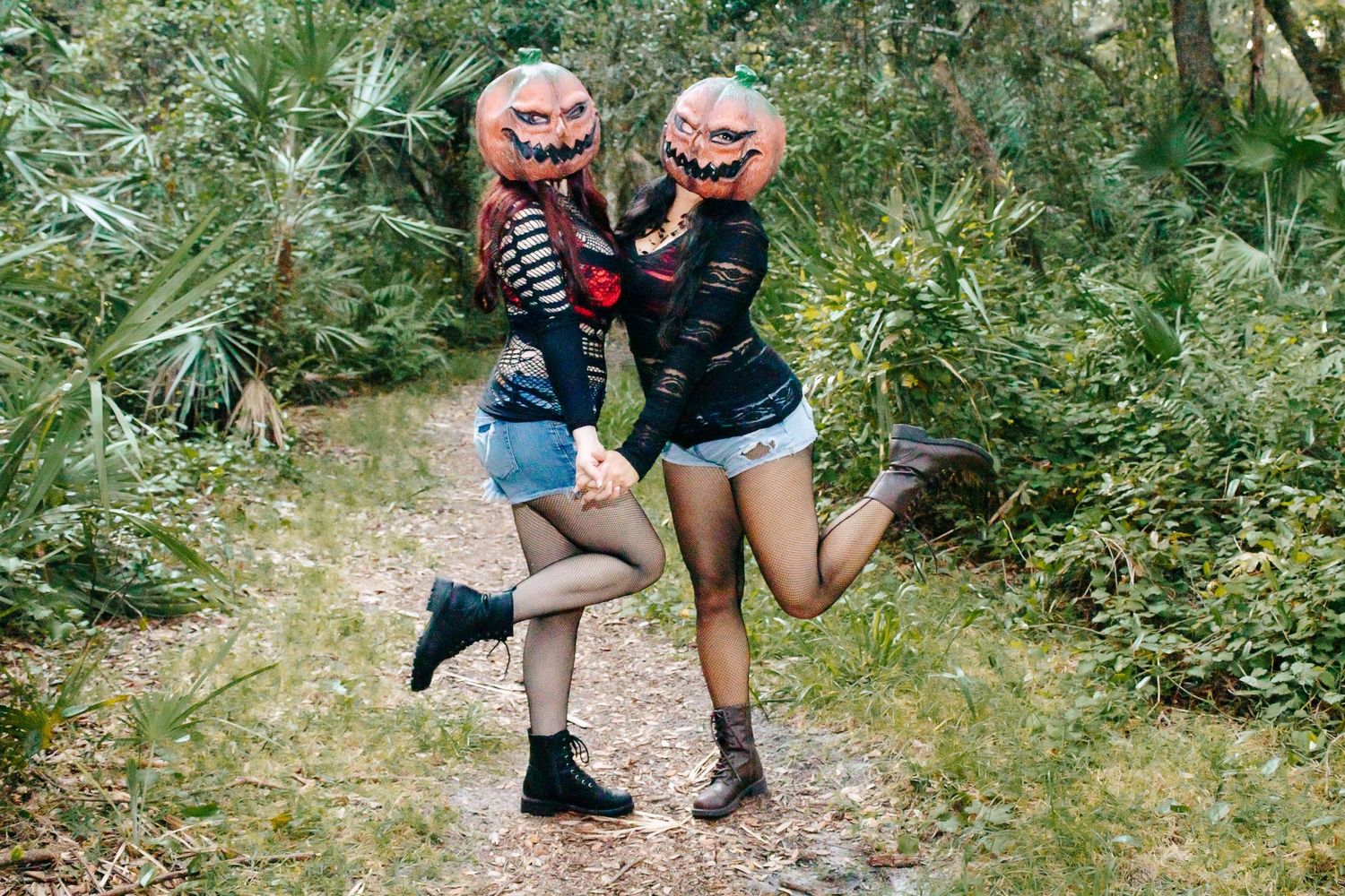 Two people in pumpkin masks and casual clothing jump together on a woodland trail.