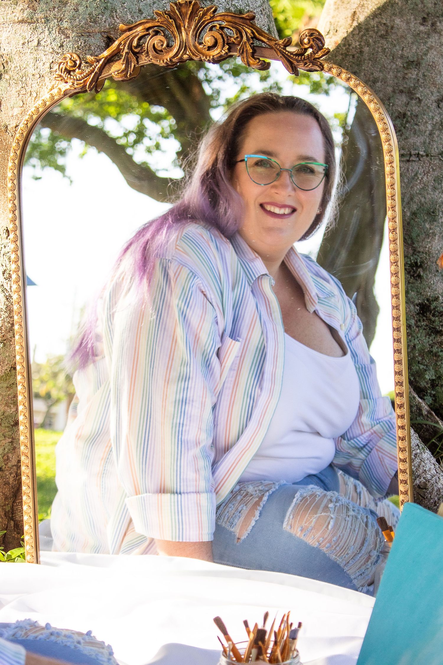 Artist relaxing outdoors with paint supplies on a white blanket under a decorative gold mirror in a garden setting.
