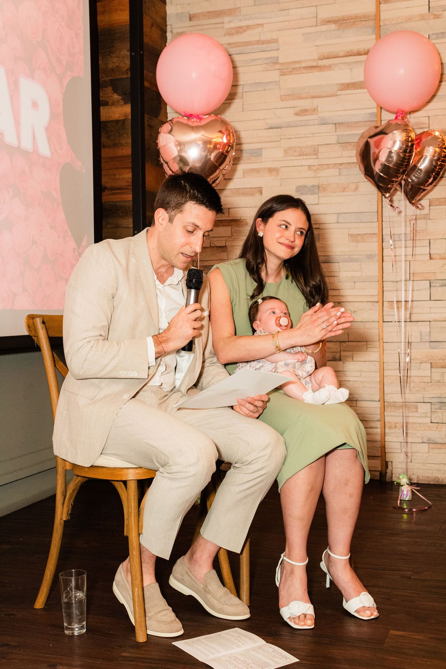 A couple seated with a baby at an event with pink balloons and decorative lighting.