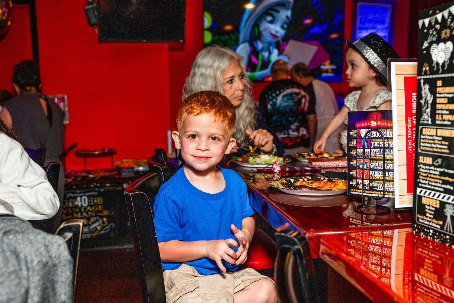 A young boy in a blue shirt sitting at a table in a restaurant or arcade.