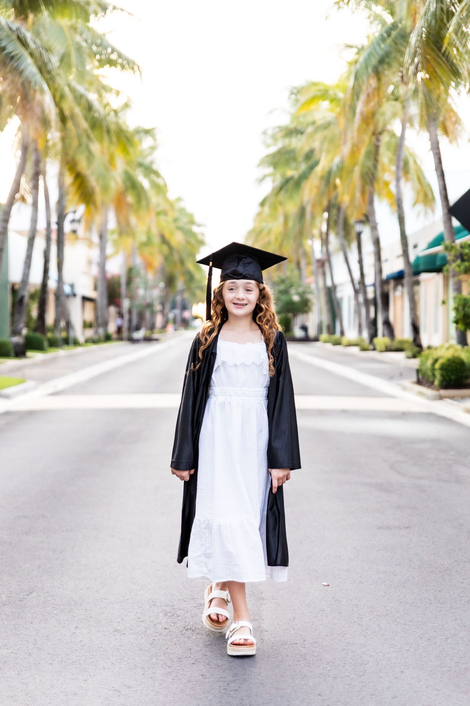 Young girl in black graduation cap and gown over white dress stands on palm-lined street, smiling proudly.