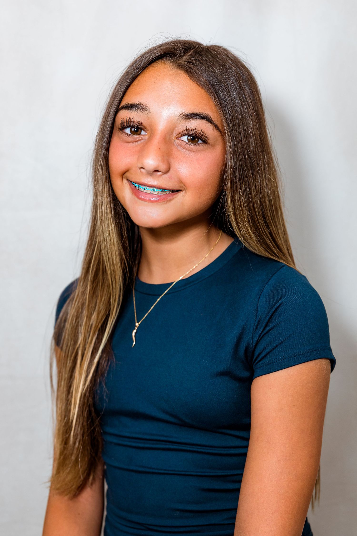 Smiling teenage girl with long brown hair, braces, and a teal shirt wearing a delicate gold necklace.