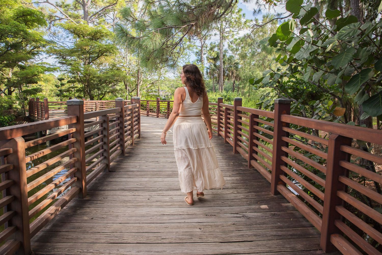Person in a flowing dress walks along a wooden boardwalk surrounded by lush green trees in dappled sunlight.