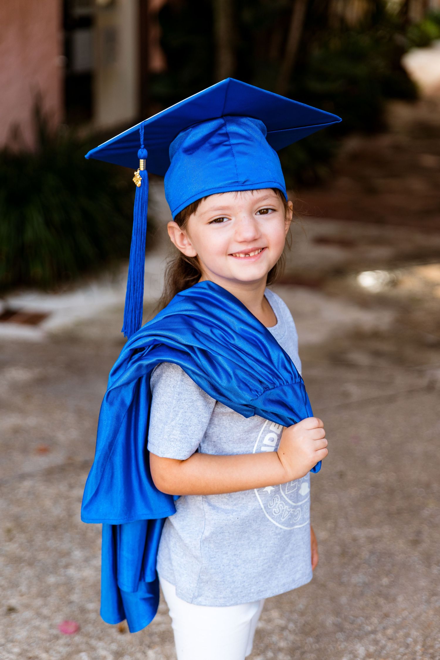 A young child wearing a gray t-shirt playfully poses with a bright blue graduation cap and gown against a blurred background.