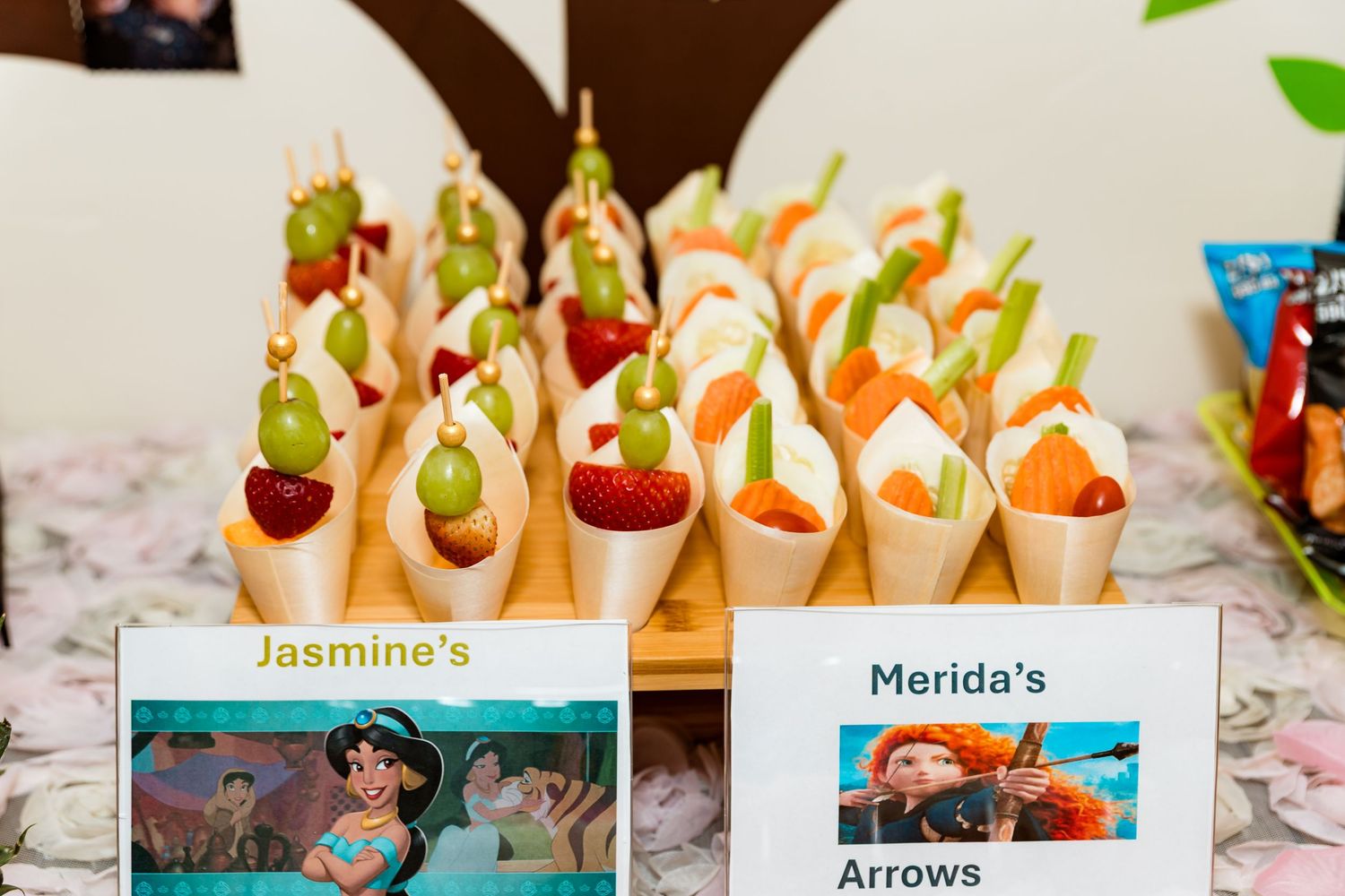 Themed party snacks with name cards displaying Jasmine and Merida's arrows on a decorated table.