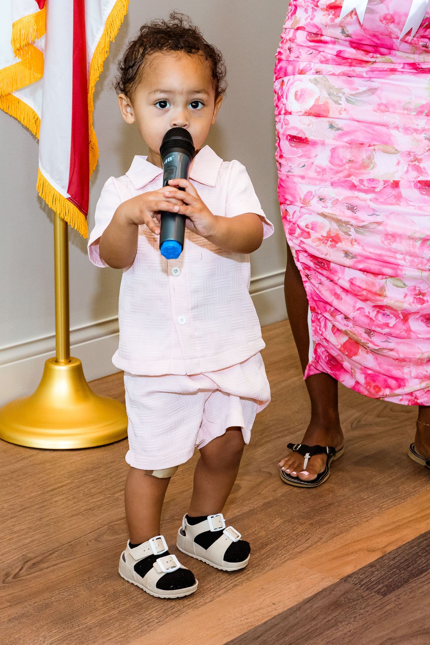 Small child in white outfit holding a microphone stands near pink decorations.