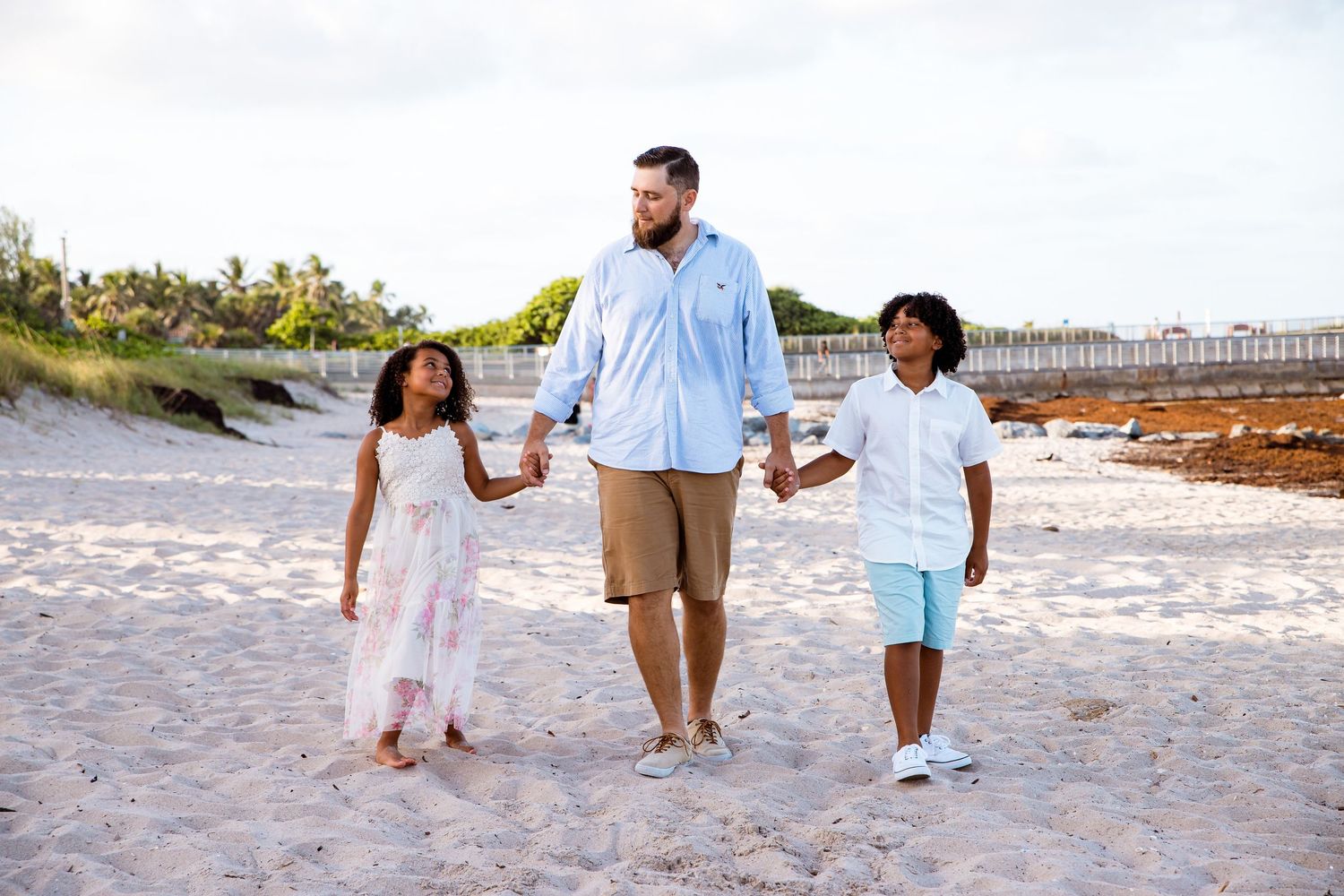 Family members walk along a scenic beach shoreline at sunset wearing coordinated white and neutral outfits.