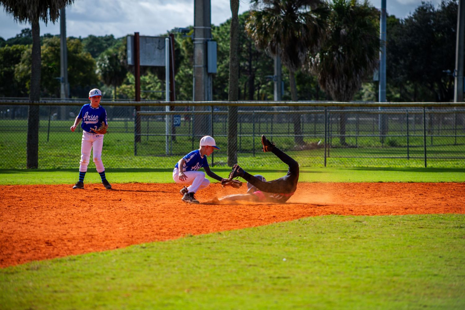 Baseball player slides into base during youth league game on orange clay diamond under blue sky and palm trees.