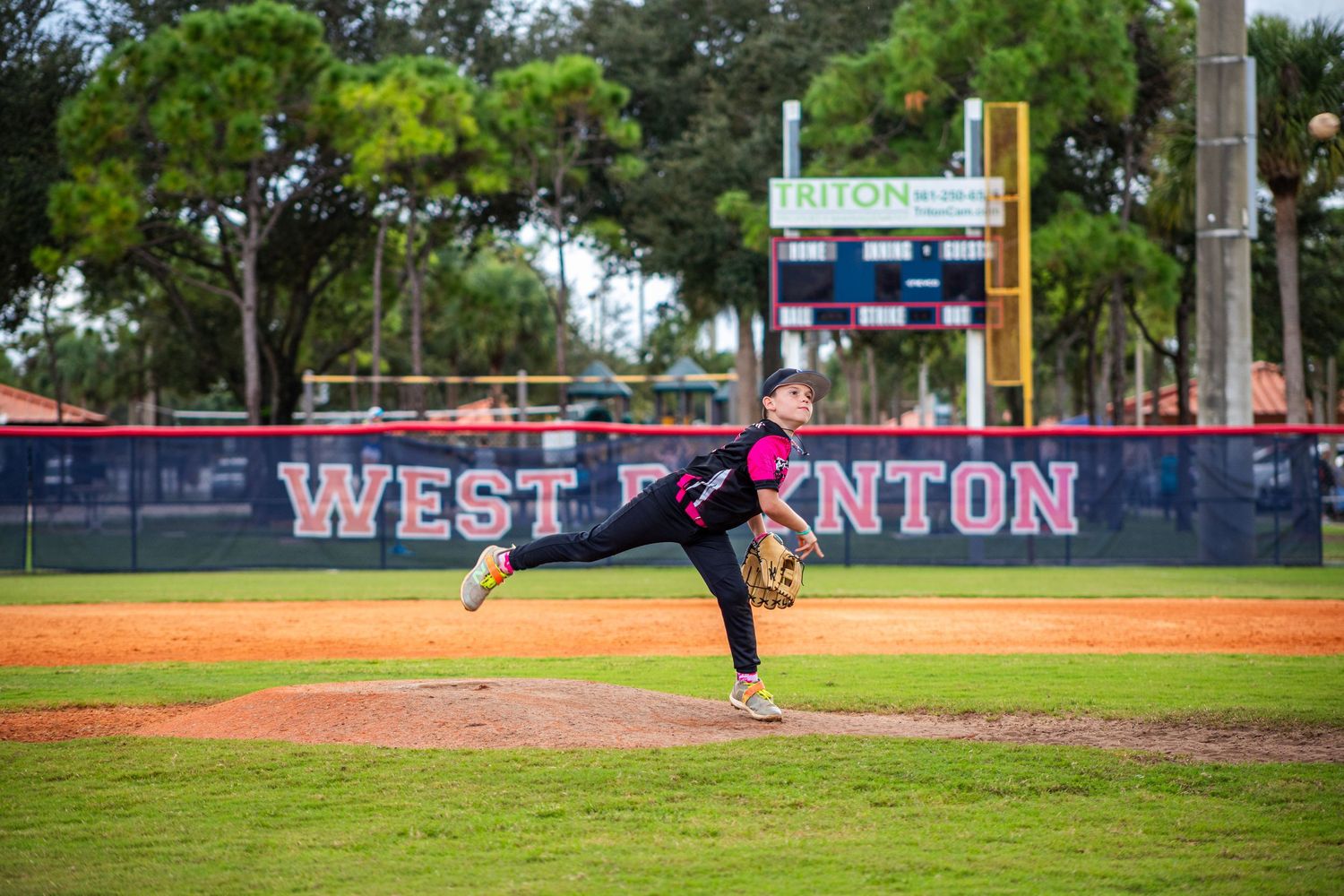 Baseball diamond with outfield fence and trees visible in the background during a game.