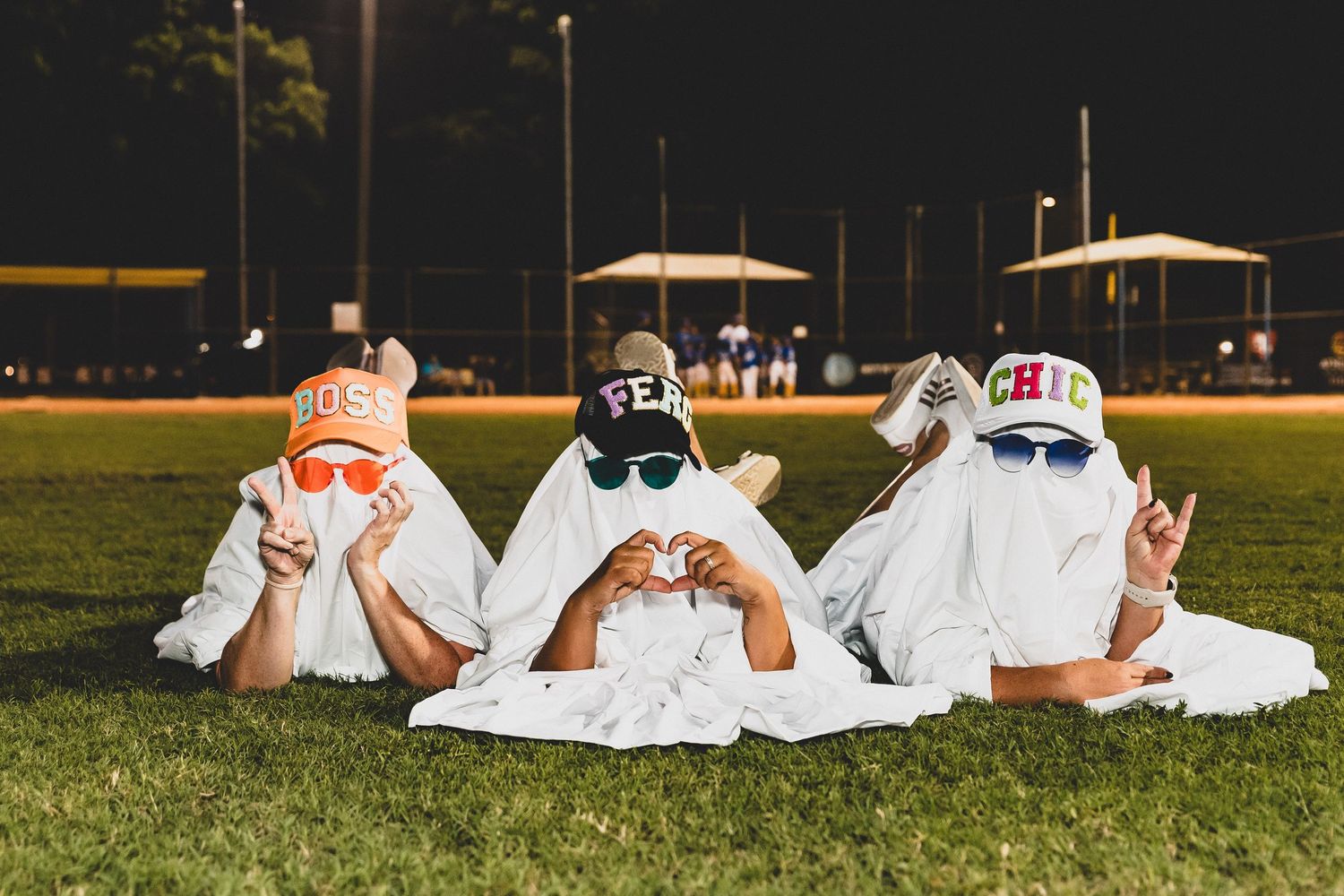 Group in ghost costumes lay on grass making playful poses under night sky.